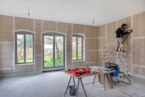Handwerker schleift Wand in renoviertem Innenraum; Arbeitsmaterialien auf Tisch im Vordergrund.