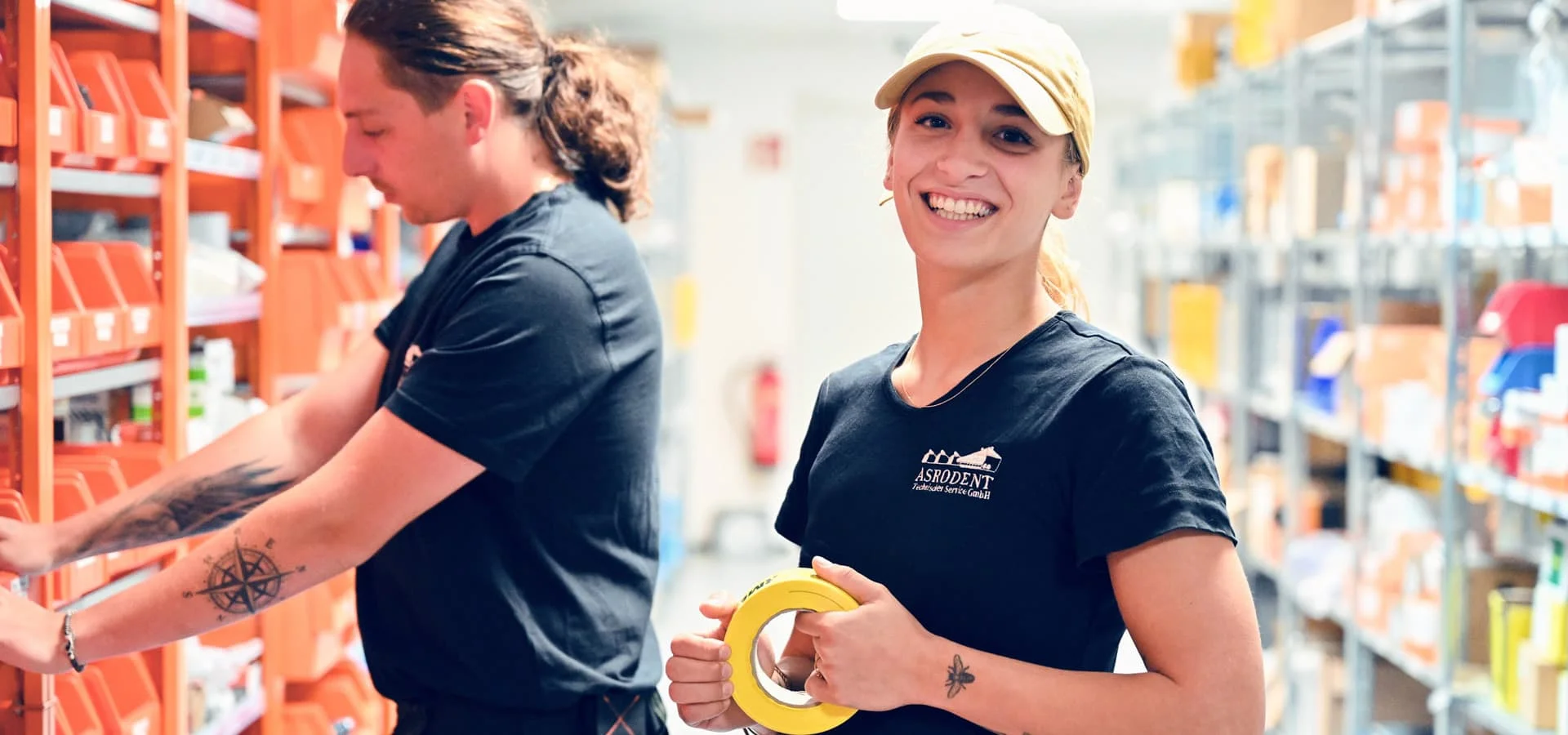 Lächelnde Lagerarbeiterin mit gelbem Klebeband in der Hand, während ein Kollege Regale im Lager bestückt.